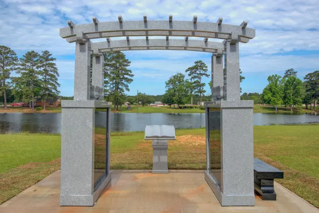 Stone memorial arch and plaque by a lakeside with trees and blue sky