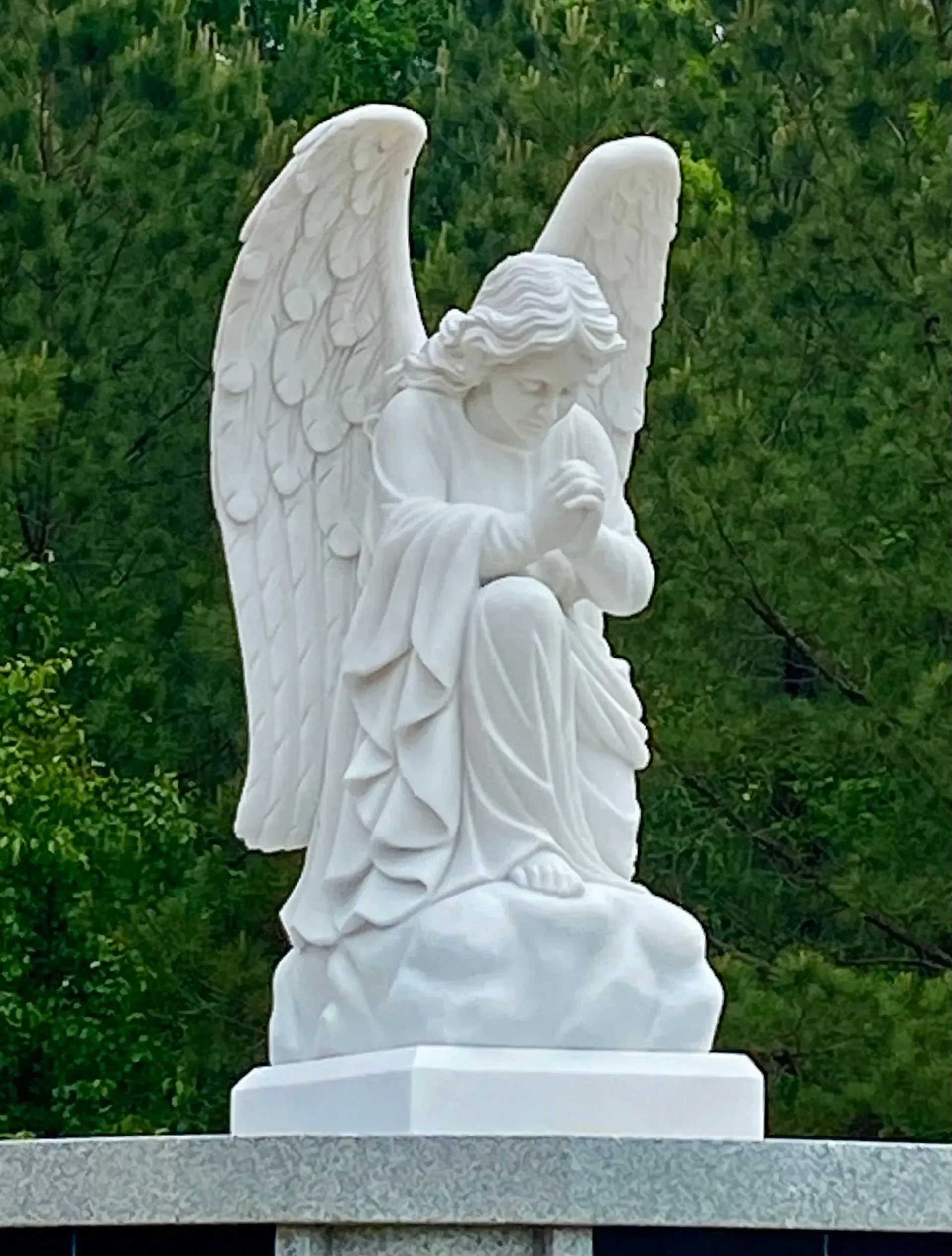 White angel statue with folded hands on a stone pedestal against green trees