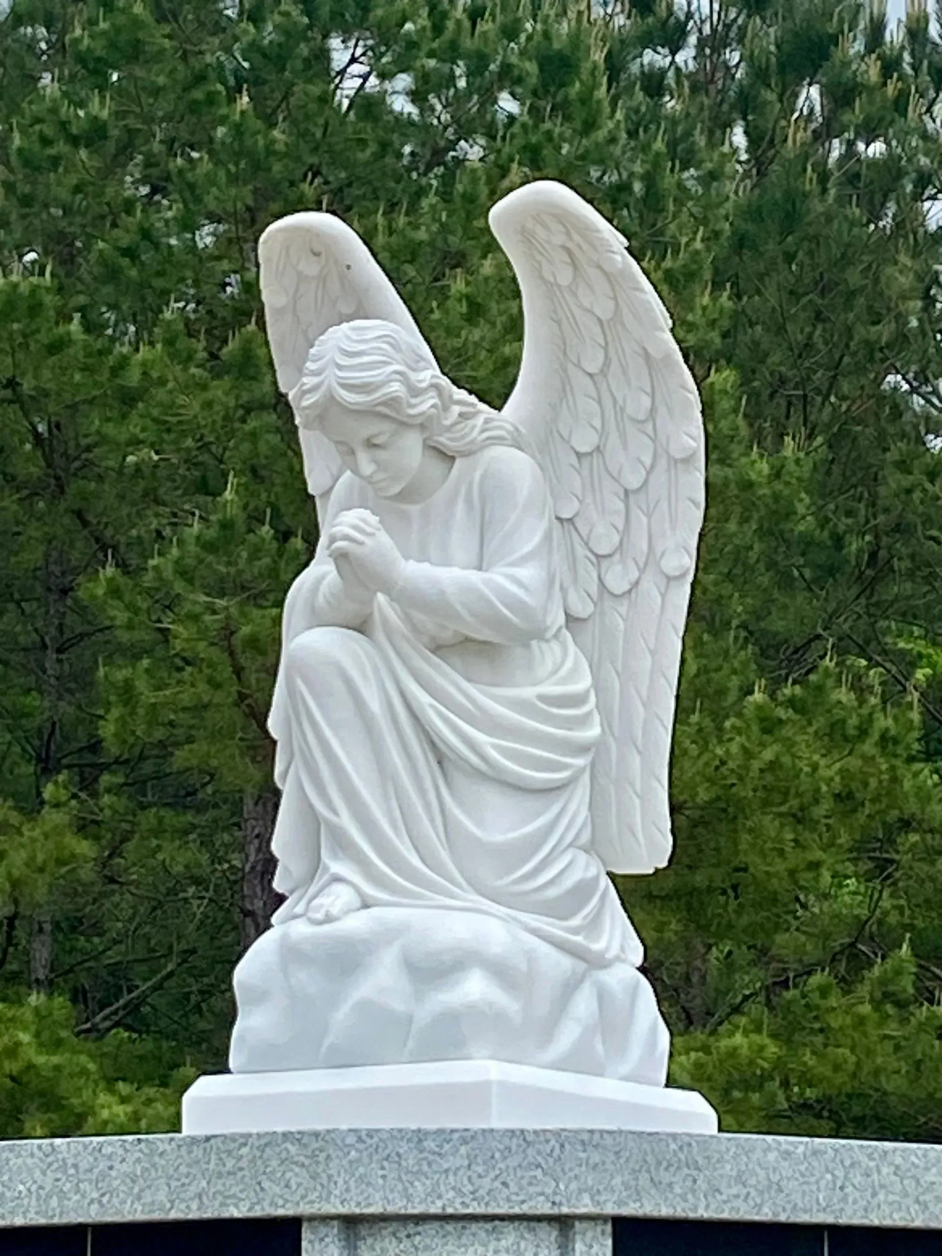White angel statue with folded wings standing on a pedestal against green trees