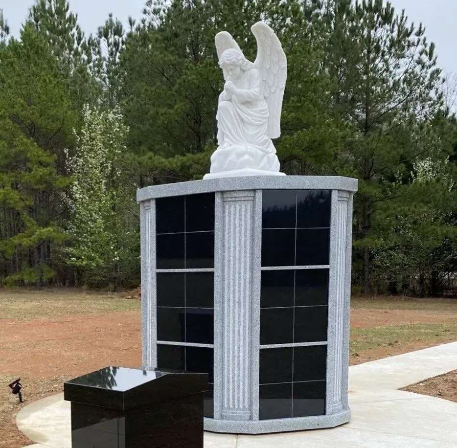 White angel statue atop a dark memorial plaque, outdoors among pine trees