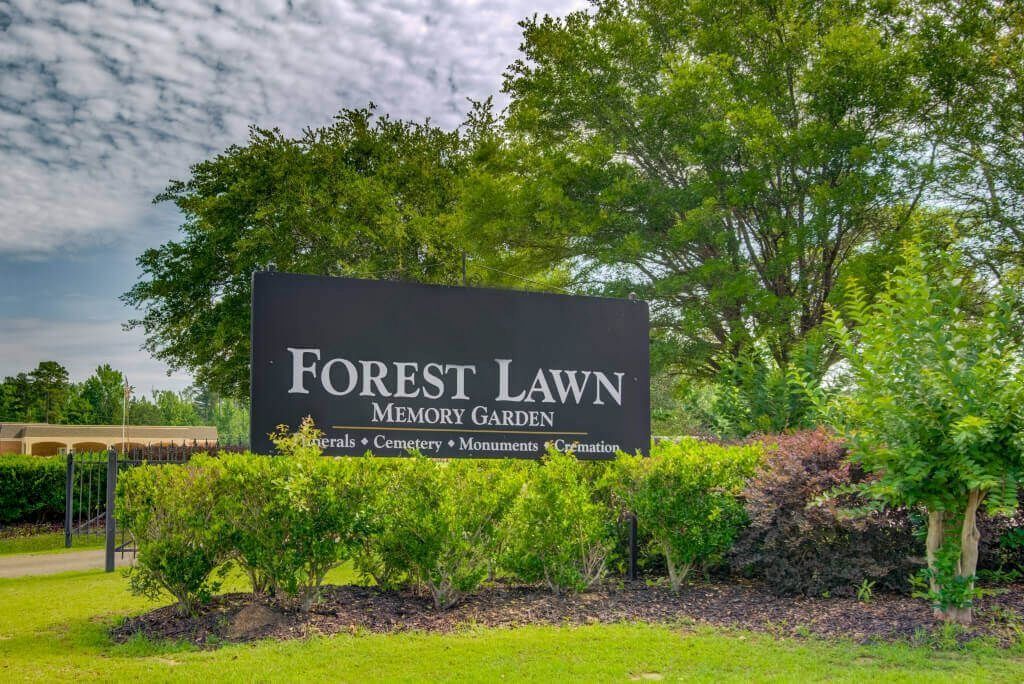 Forest Lawn Memorial Gardens sign beside lush trees and shrubs under a cloudy sky
