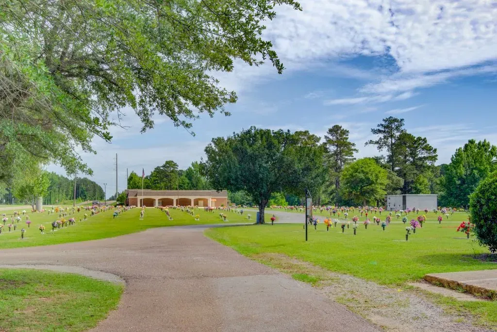Gravel path through a grassy cemetery with scattered headstones, trees, and a small brick building under a blue sky