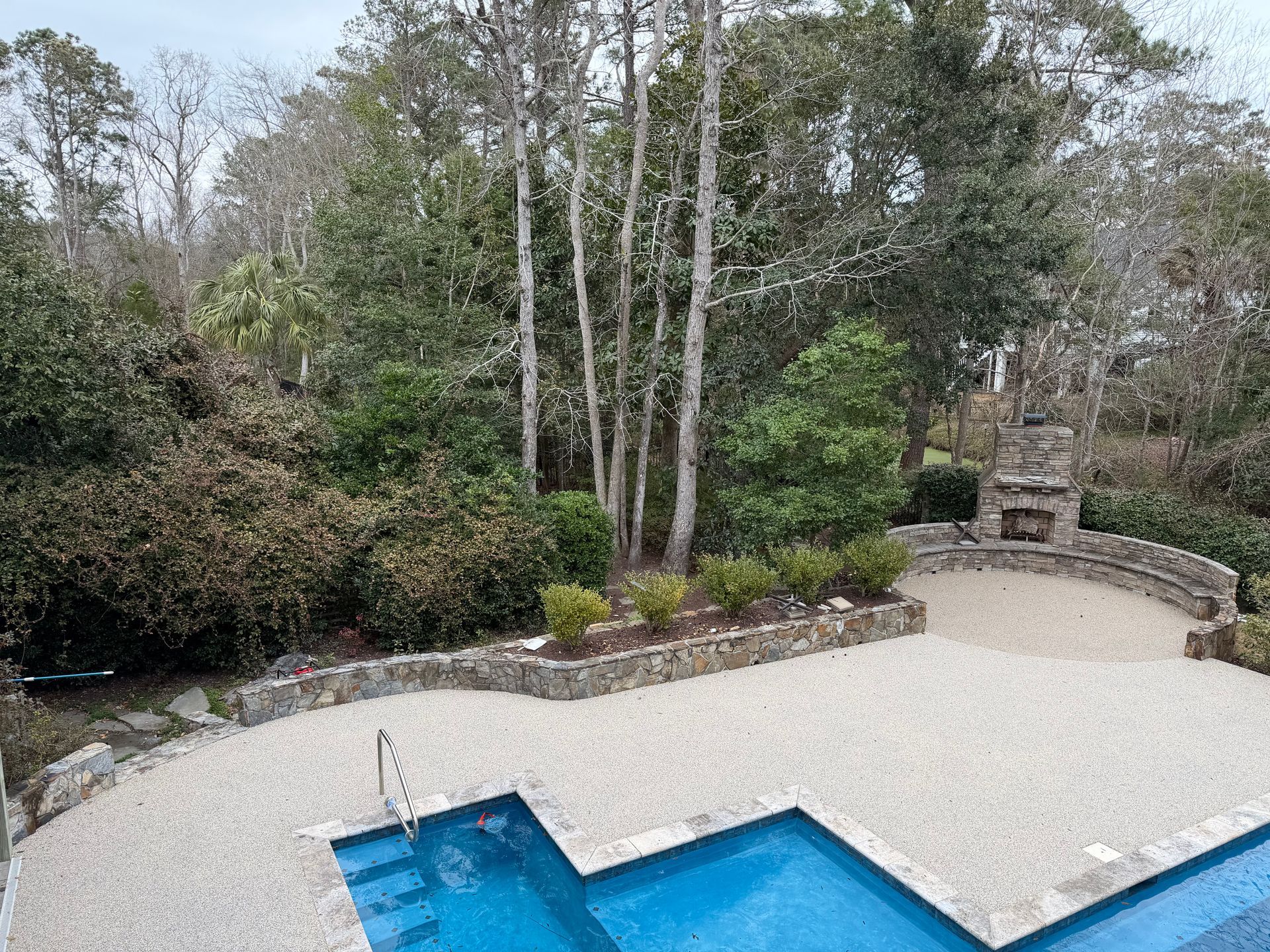 Backyard pool with blue water, white patio, curved stone wall, and wooded garden area