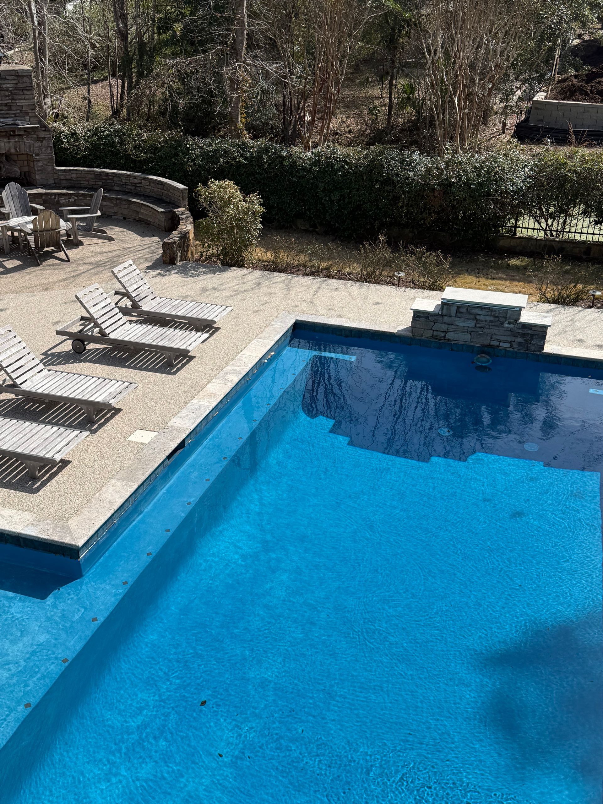 Blue swimming pool with lounge chairs on a stone patio and greenery in the background
