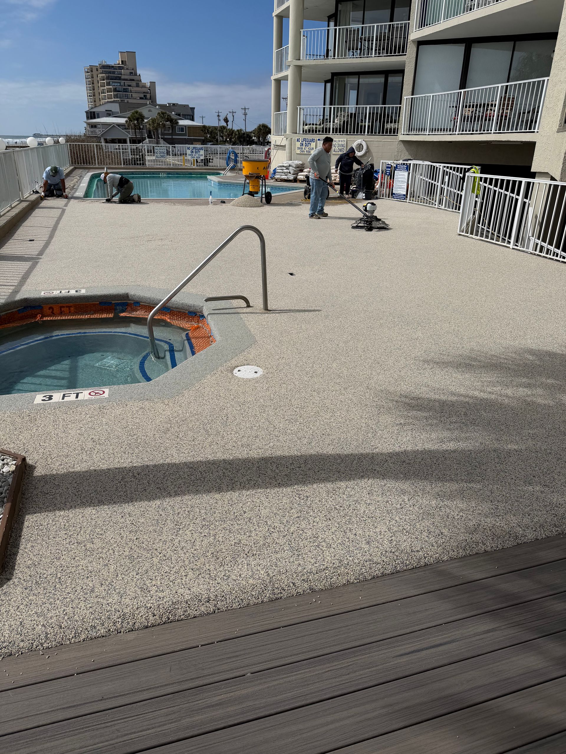 Rooftop pool deck with a hot tub, lounge chairs, and people near a tall modern building by the water.