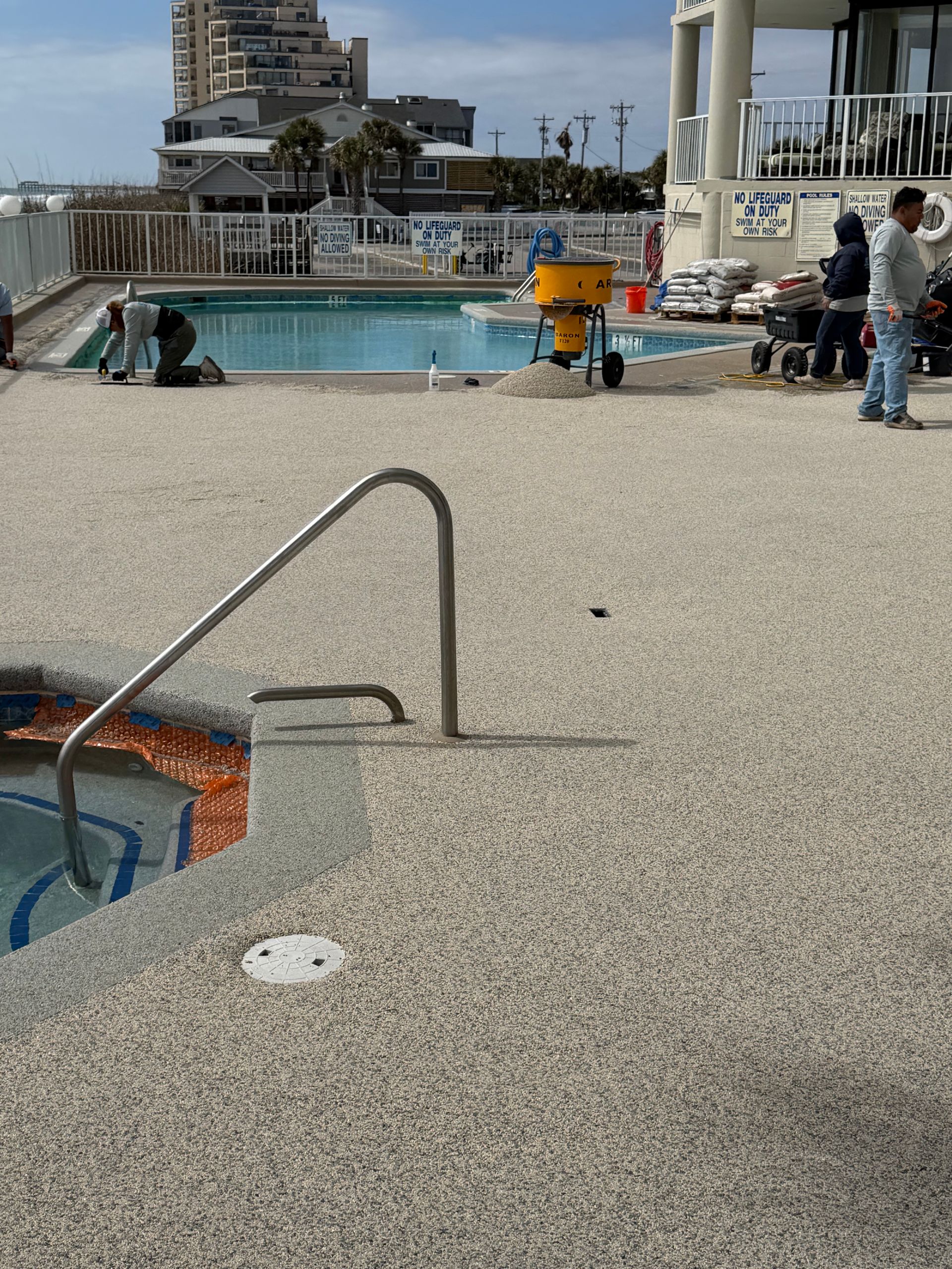 Outdoor pool deck with a handrail, lounge chairs, and people near the water under a cloudy sky