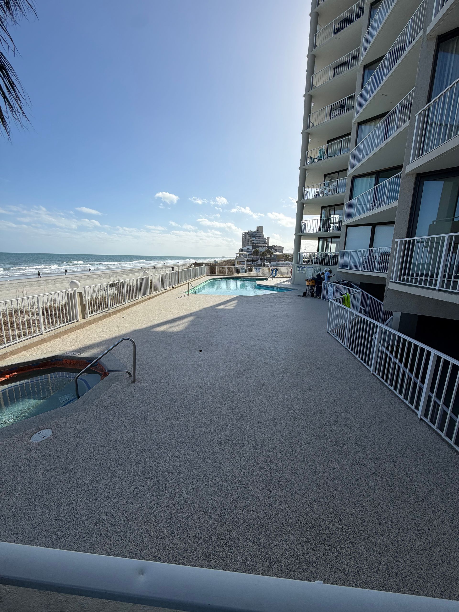 Beachfront pool beside a high-rise hotel, with lounge chairs, railing, and ocean view under a blue sky