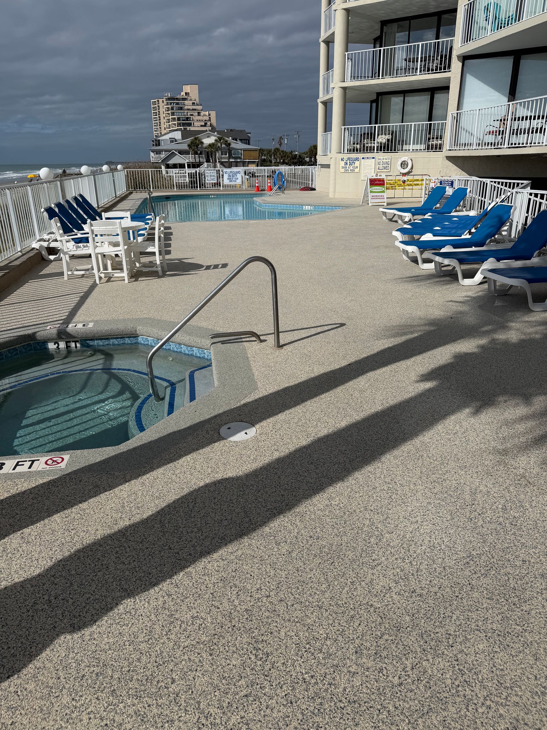 Rooftop pool deck with blue lounge chairs, hot tubs, and ocean view under cloudy skies
