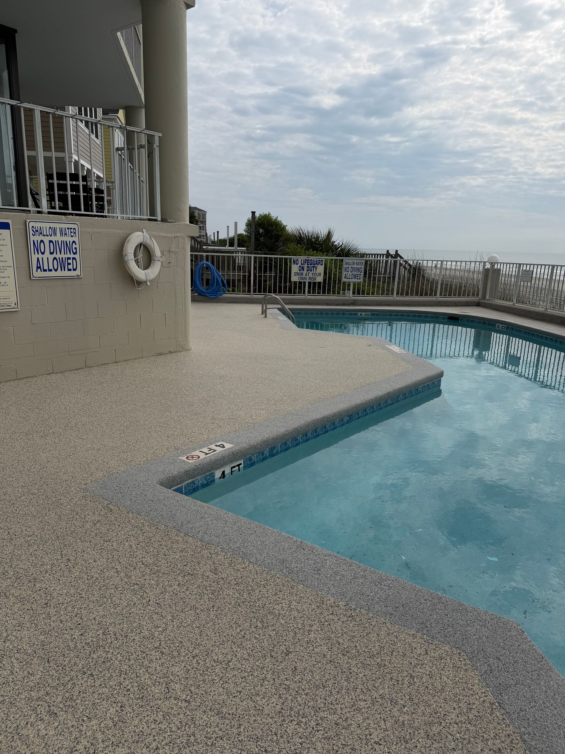 Poolside deck and curved swimming pool beside a building, overlooking the ocean under a cloudy sky