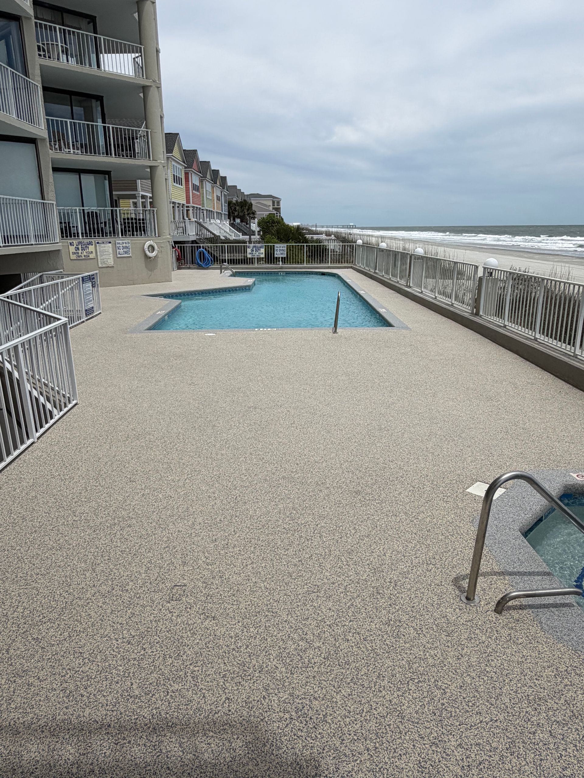 Oceanfront pool on a concrete terrace beside the beach, with waves and overcast sky in the background