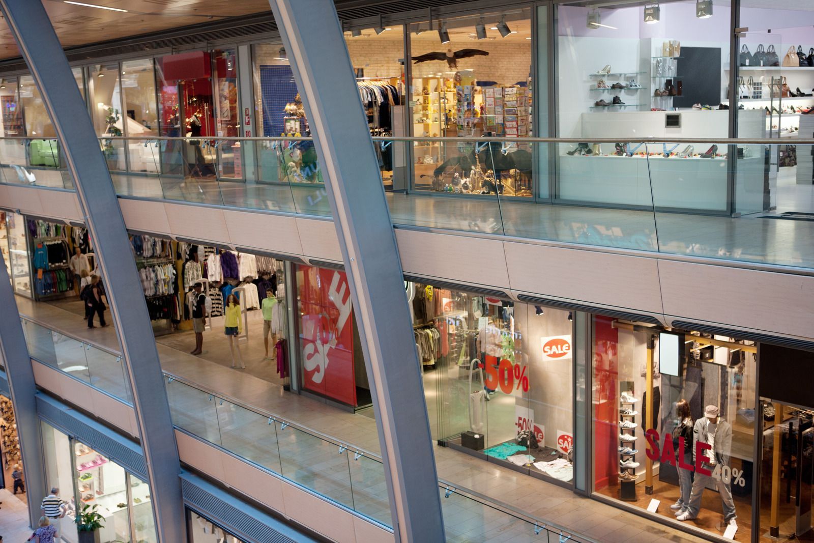 Interior view of a modern shopping mall with glass railings, storefronts, and people walking on multiple levels