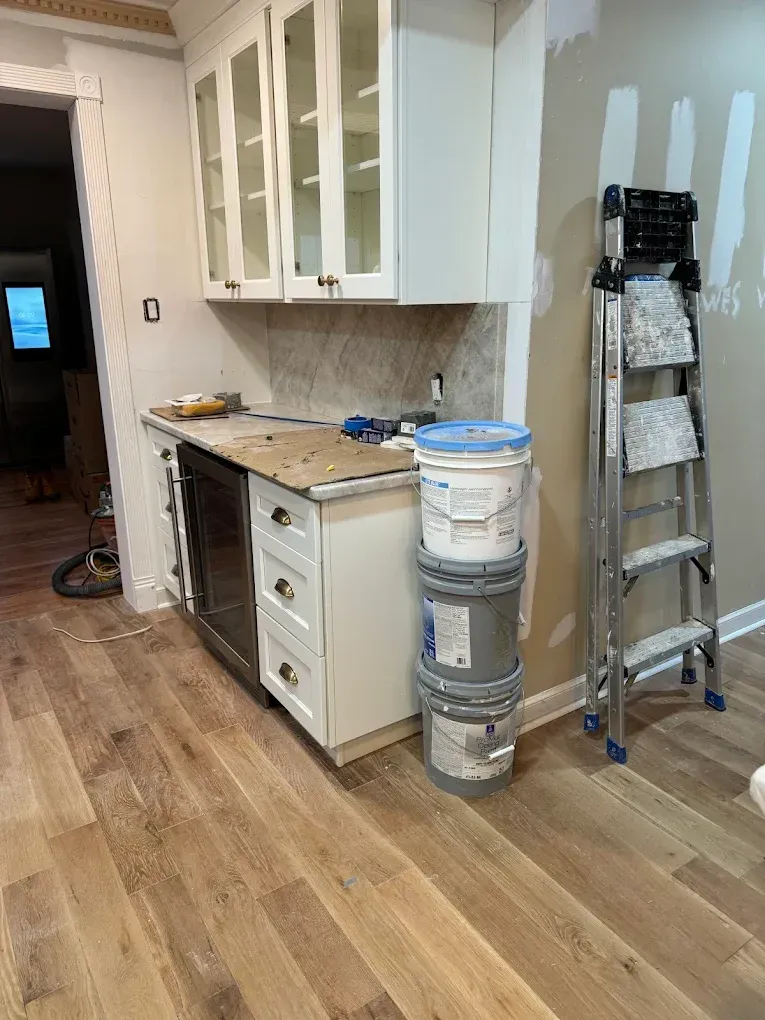 Kitchen under renovation with cabinets, paint buckets, ladder, and wood flooring.
