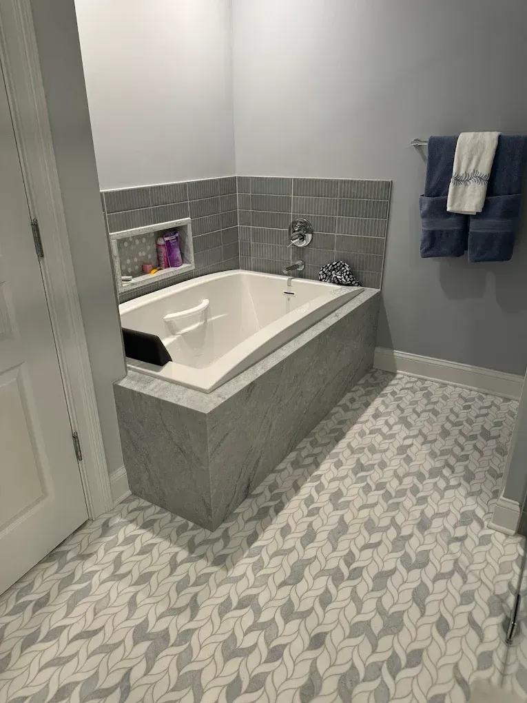 Bathroom with a tub surrounded by gray tile, and patterned tile flooring. Towel rack with blue towels.