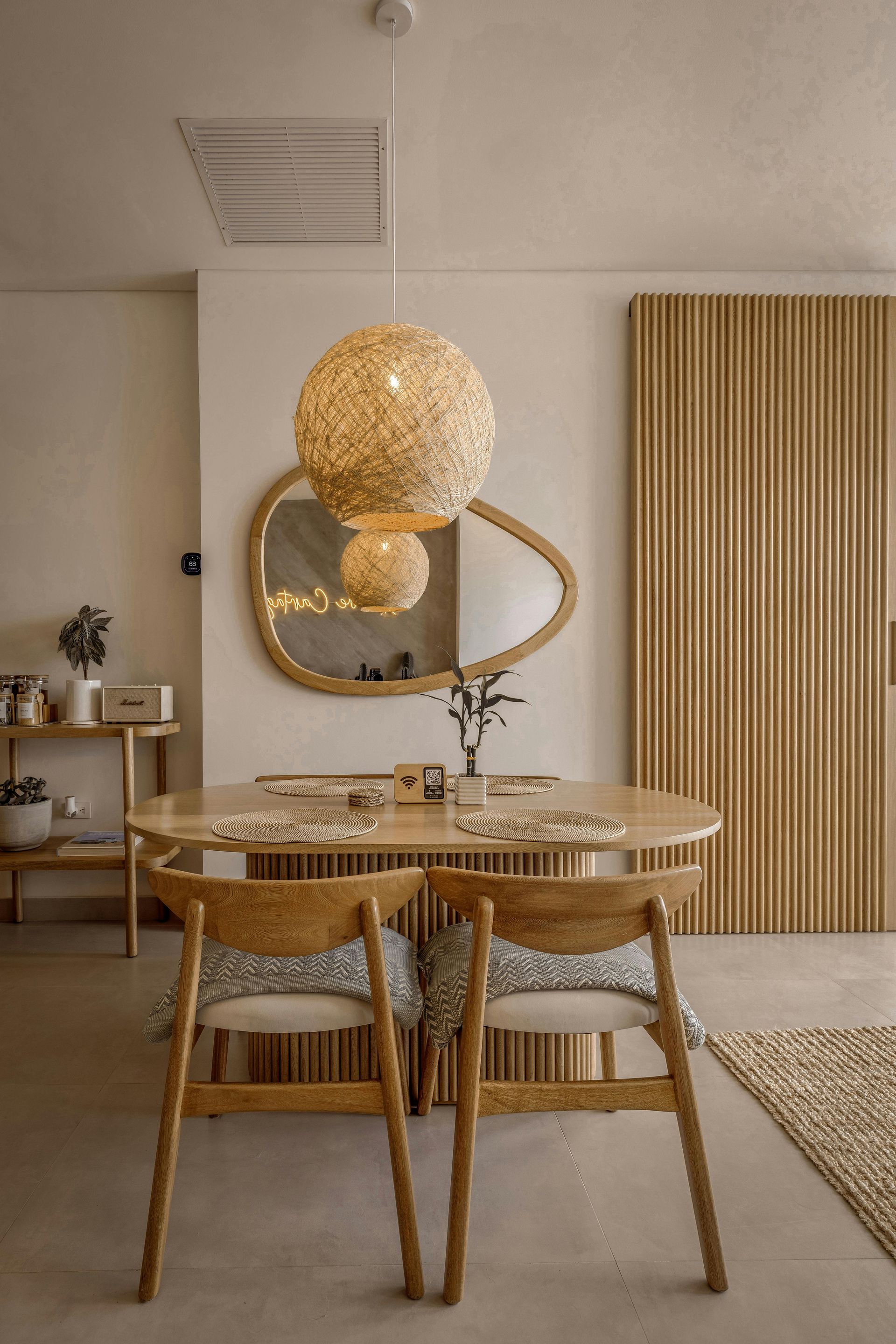 A dining area with a wooden table, two chairs, a textured pendant light, and an organic-shaped mirror on a beige wall.