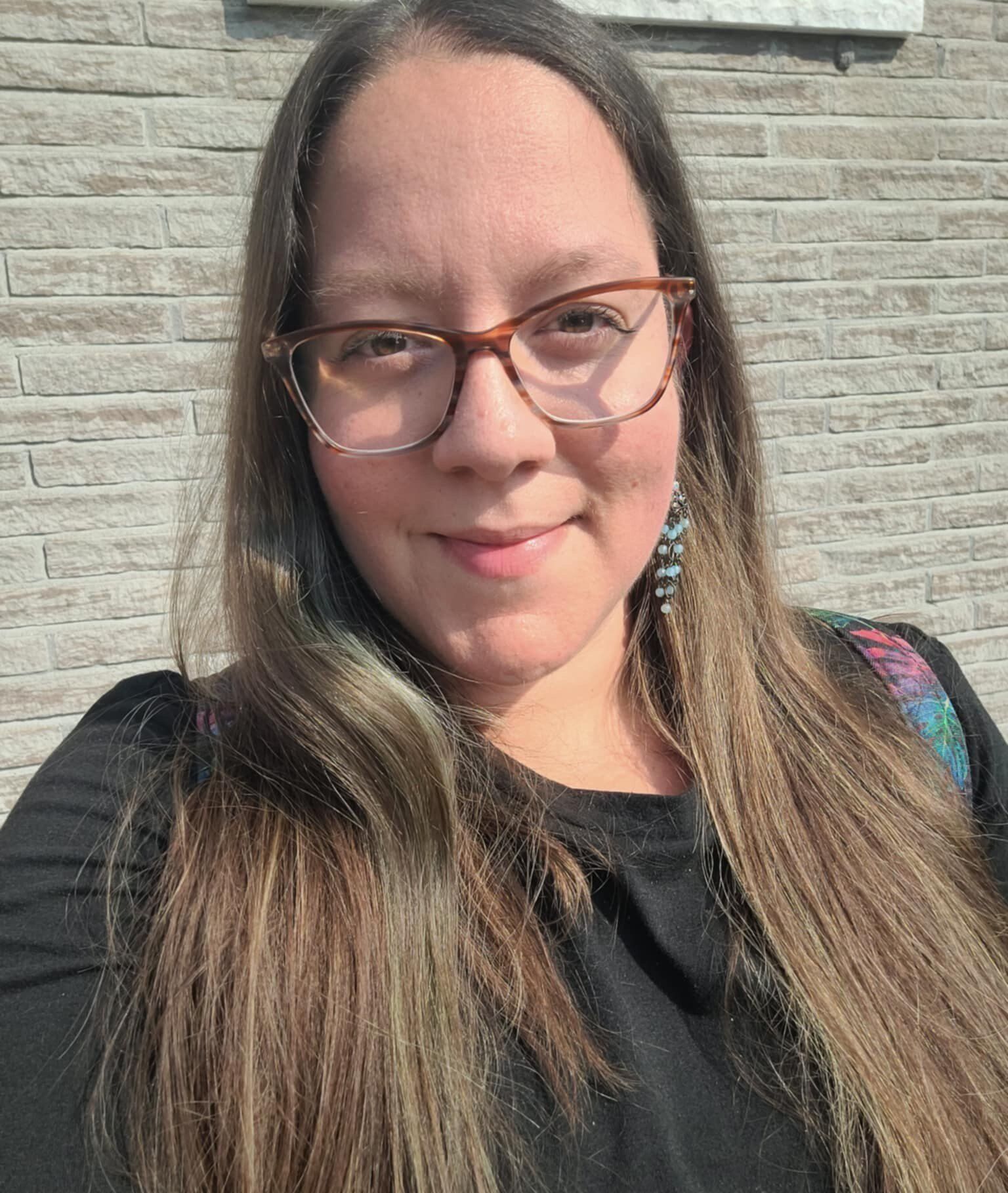 Femme aux longs cheveux bruns et portant des lunettes, souriant à l'extérieur devant un mur de pierre.