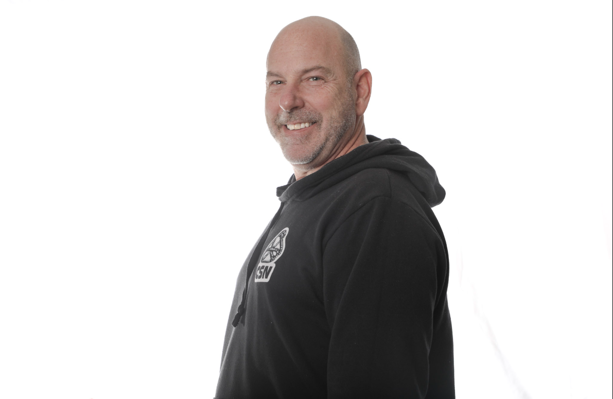 Un homme chauve souriant, vêtu d'un sweat à capuche noir, se détache sur un fond blanc.