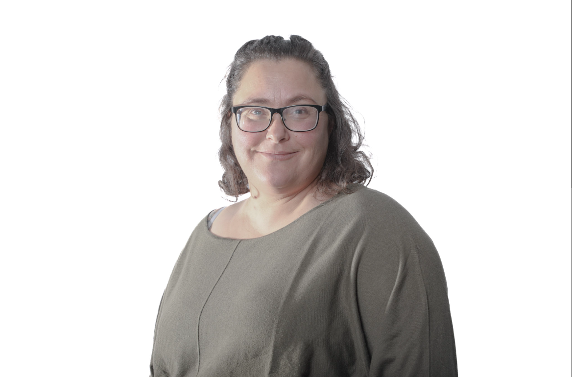 Personne portant des lunettes et un haut olive, souriant sur fond blanc