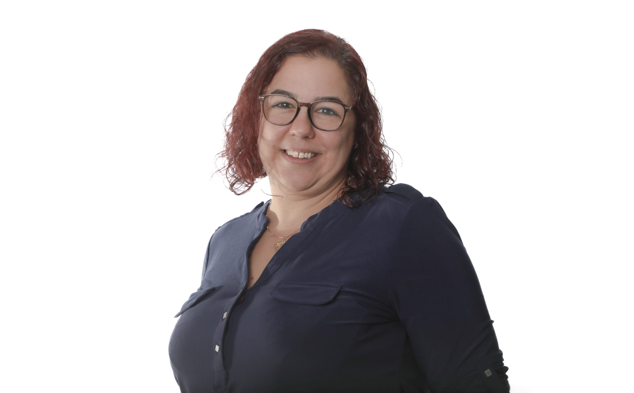 Femme à lunettes et aux cheveux roux bouclés, souriante dans un chemisier bleu marine sur fond blanc