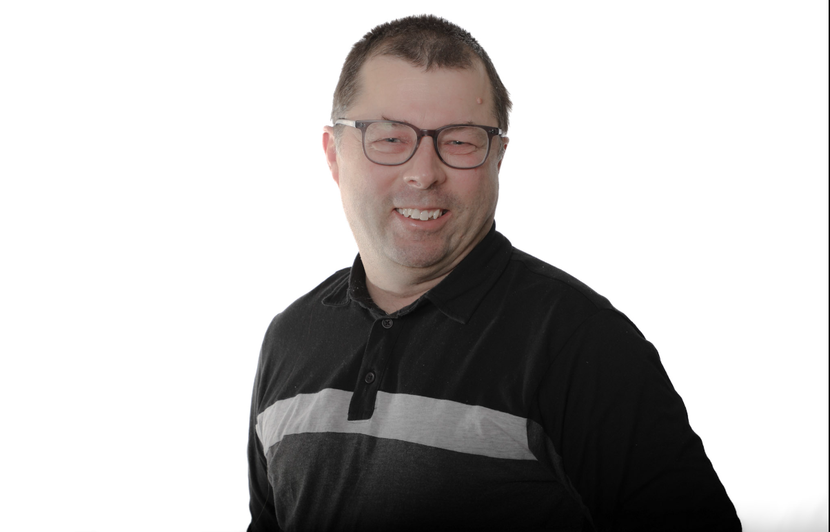 Homme souriant en chemise noire à rayures grises, portant des lunettes, sur fond blanc