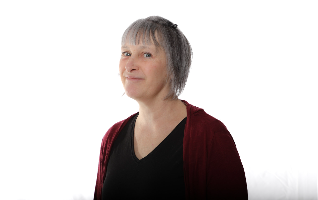 Personne aux cheveux gris courts portant un haut noir et un cardigan bordeaux sur fond blanc