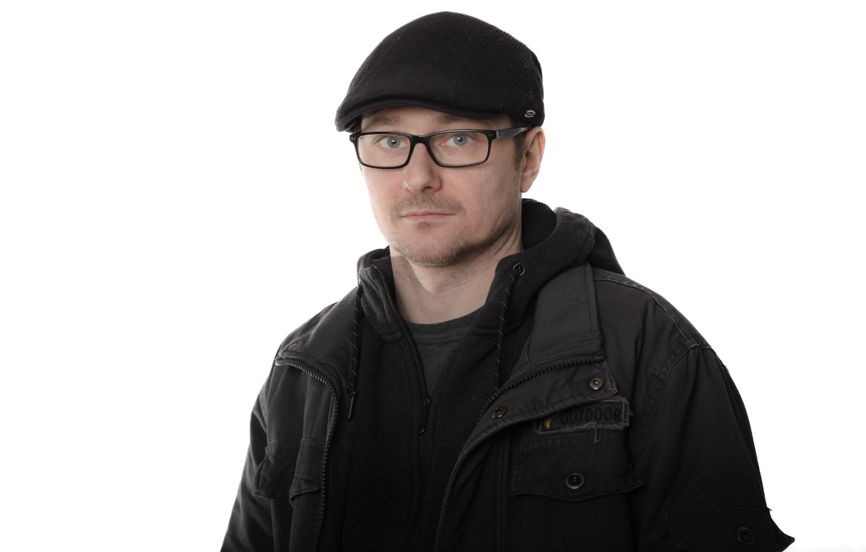 Homme portant une casquette noire, des lunettes et une veste sur fond blanc