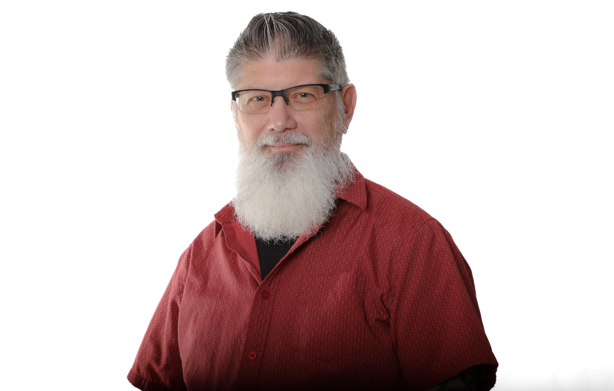 Homme portant des lunettes et une longue barbe blanche, vêtu d'une chemise rouge sur fond blanc