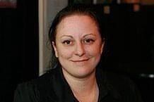 Portrait d'une personne esquissant un sourire, vêtue d'un haut noir sur fond sombre.