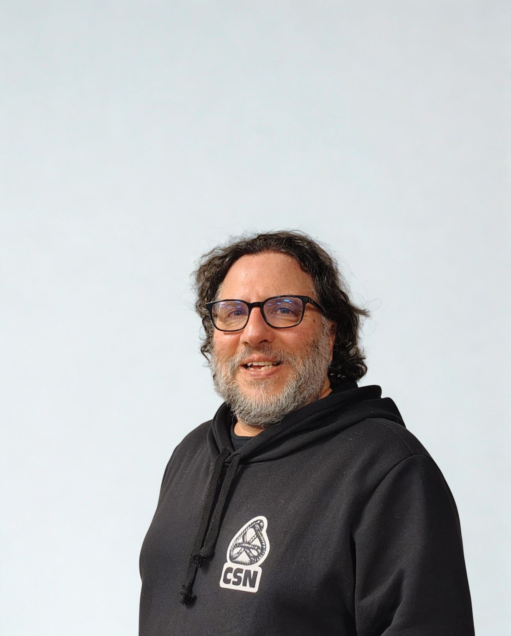 Personne portant des lunettes et des cheveux bouclés, vêtue d'un sweat à capuche foncé, souriant sur un fond gris clair