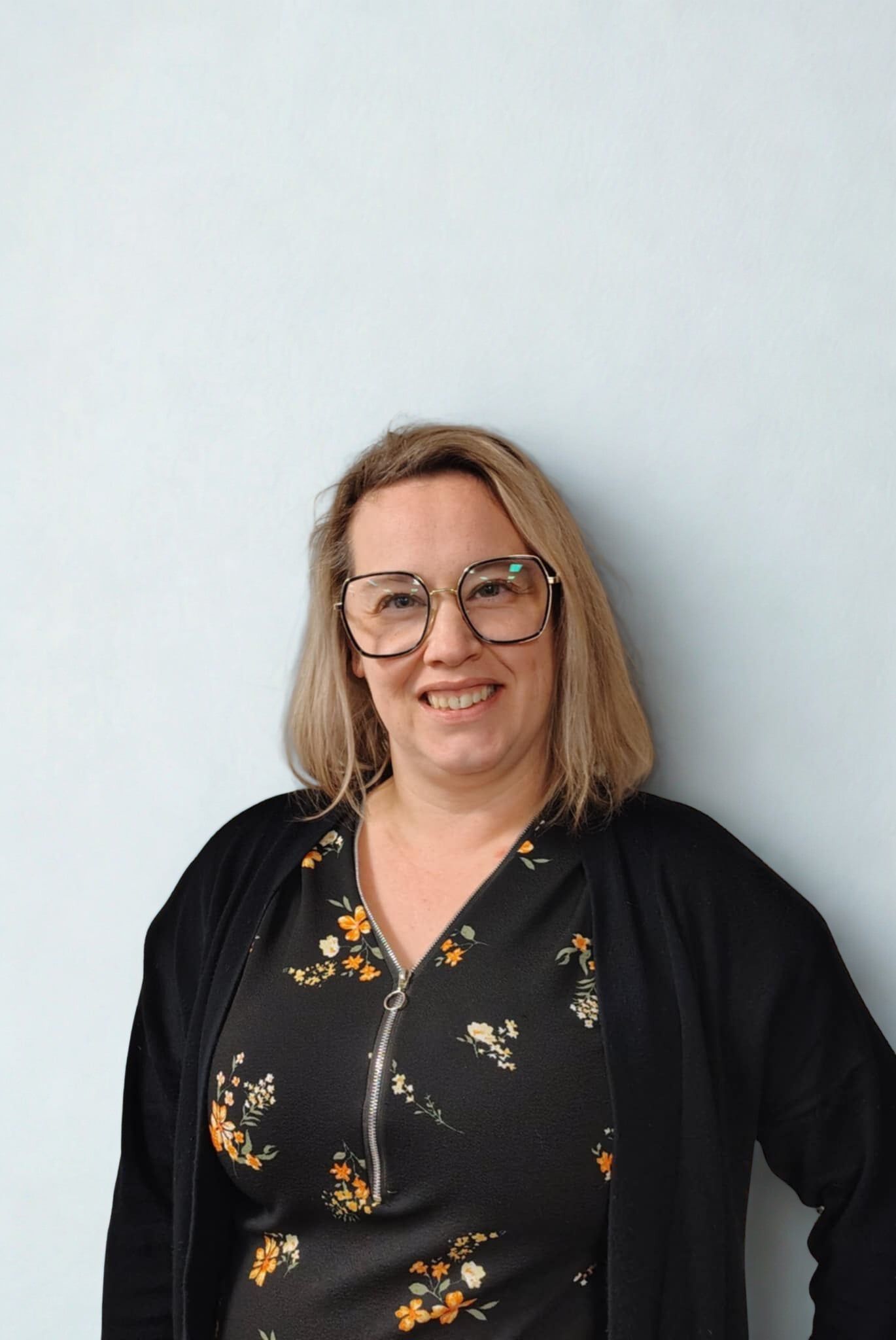 Femme à lunettes souriant devant un mur clair et uni, vêtue d'un haut à fleurs noir et d'un cardigan.