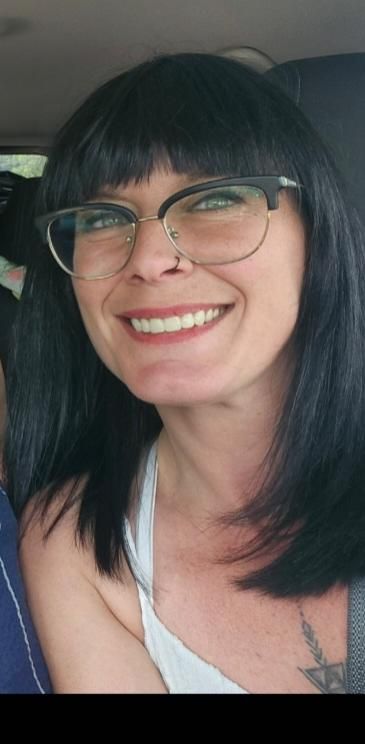 Femme souriante aux cheveux noirs et portant des lunettes, assise dans une voiture et vêtue d'un débardeur blanc.