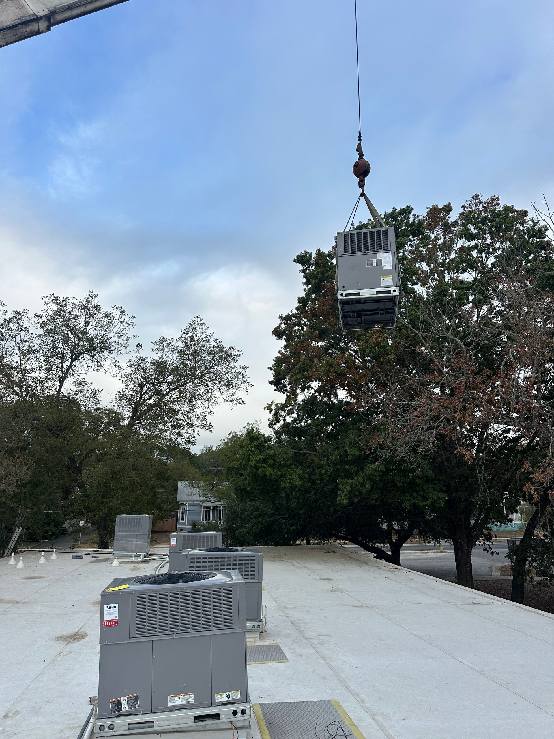 A crane lifting an HVAC unit onto a flat rooftop with existing units visible; trees and cloudy sky in background.