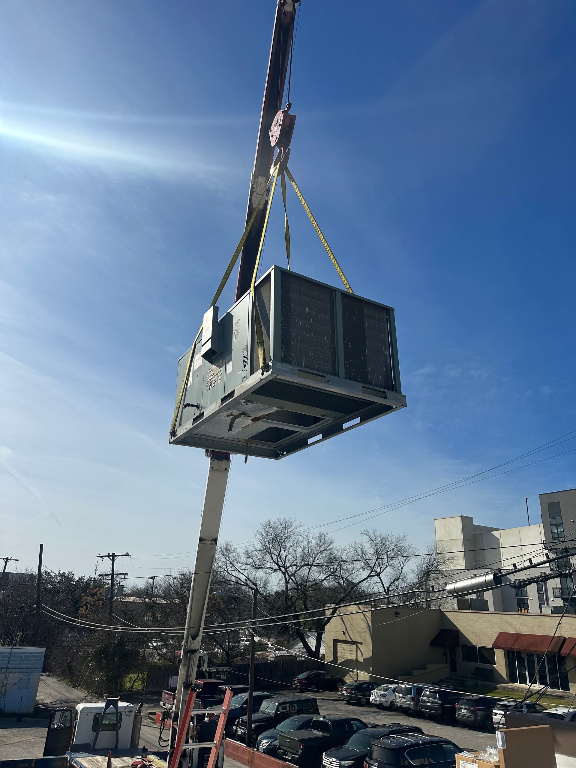 Crane lifting a large HVAC unit in front of a blue sky, trees, and buildings.