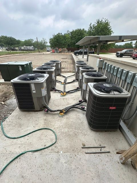 Row of air conditioning units connected by pipes on a concrete pad, outdoors.