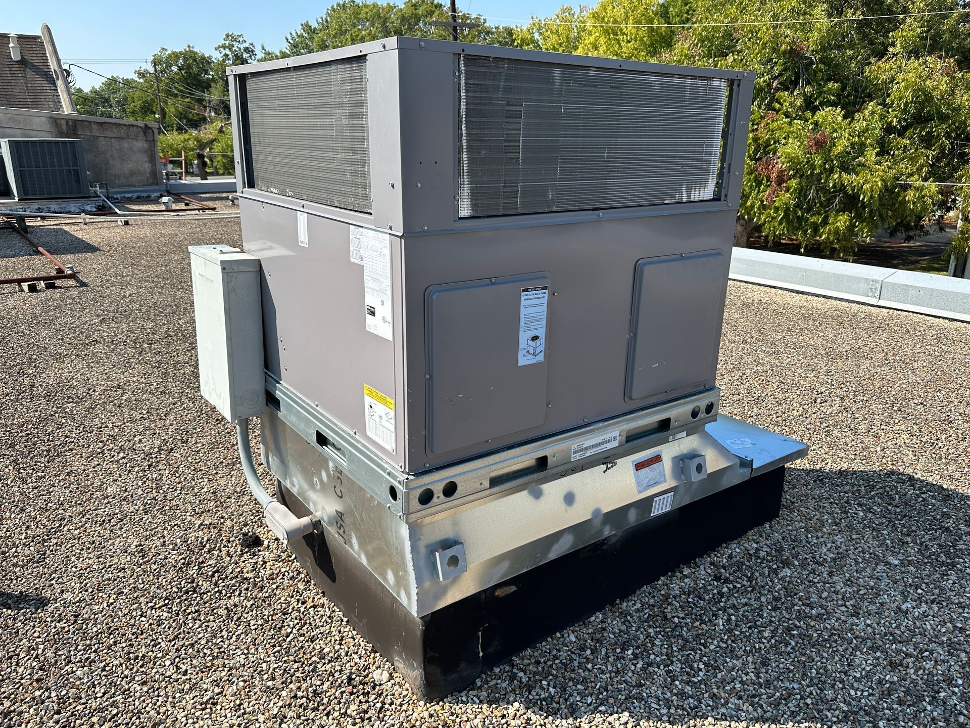 Rooftop HVAC unit on a flat roof. Gray metal box with vents and access panels. Blue sky and trees in background.