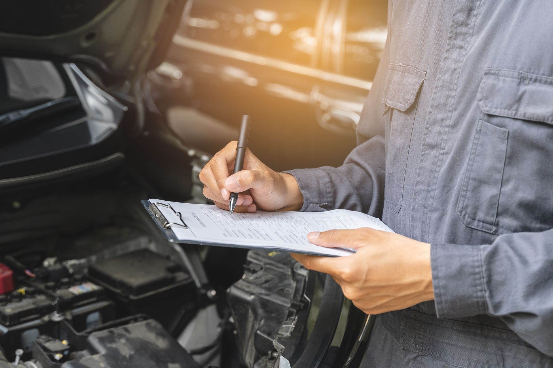 Mechanic writing on a clipboard beside an open car engine in a garage