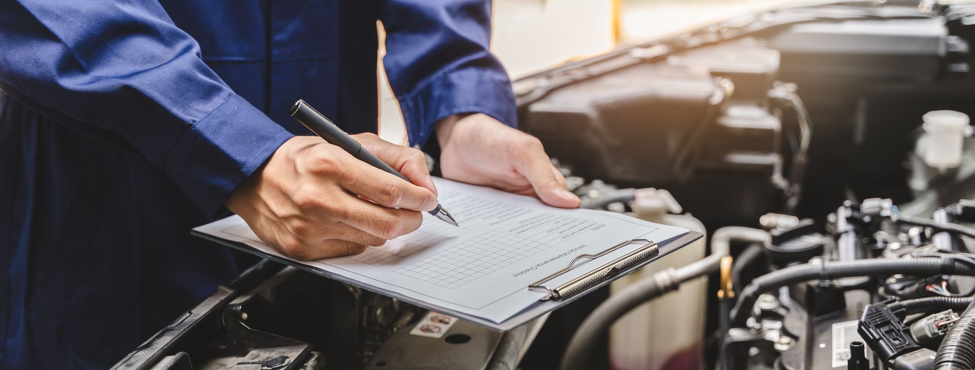Mechanic writing on a clipboard beside an open car engine in a garage
