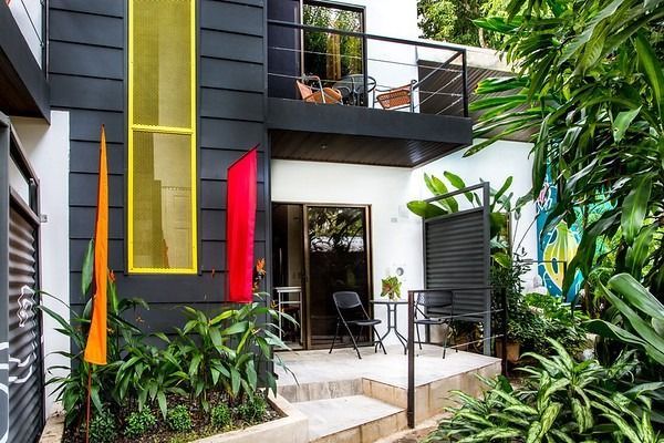 Modern house exterior with colorful hanging art, balcony, and patio surrounded by lush greenery