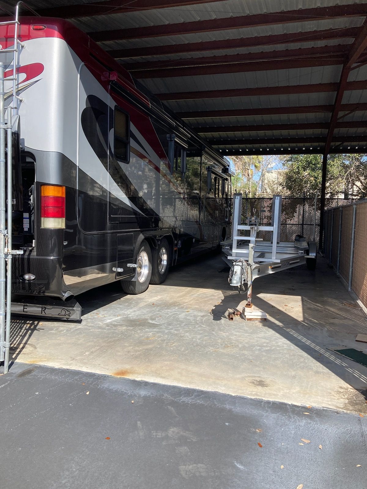 Motorhome parked under a metal carport beside a driveway and a trailer dolly.