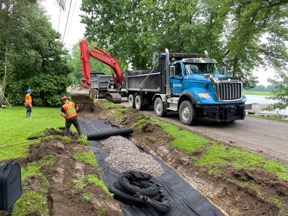 Des ouvriers et une excavatrice posent des tuyaux noirs à côté d'un camion bleu en bord de route.