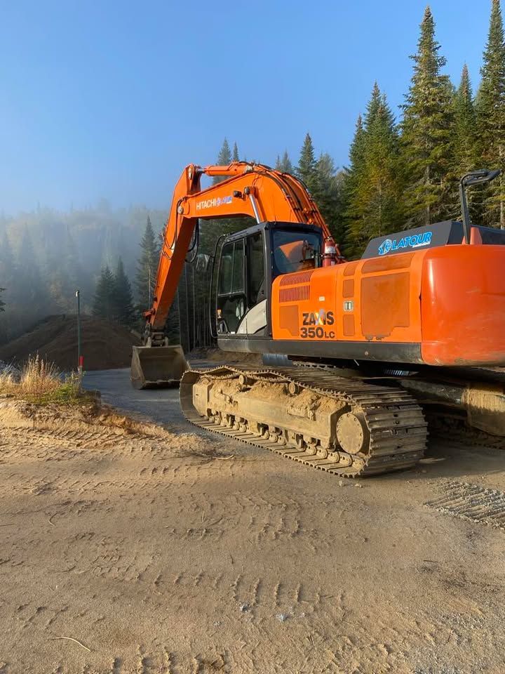 Une pelleteuse orange sur un chemin poussiéreux en bordure de forêt, avec une autre pelleteuse en arrière-plan.