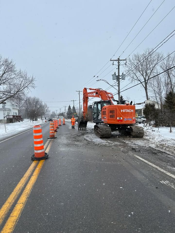 Une pelleteuse orange et des cônes de signalisation bloquent une zone de travaux en bord de route enneigée sous un ciel couvert.