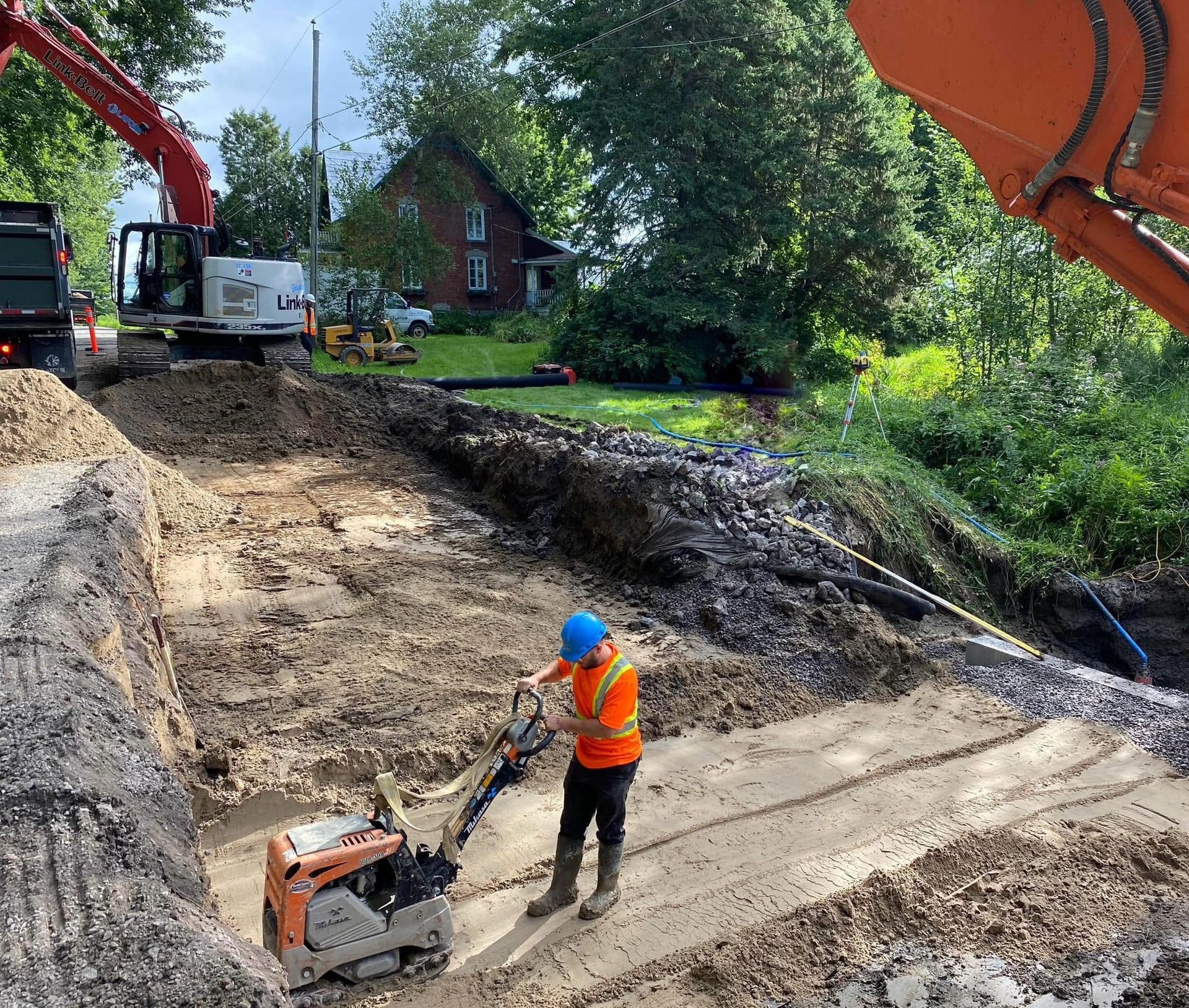 Un ouvrier du bâtiment, vêtu d'un gilet orange et d'un casque bleu, manipule une plaque vibrante sur un chantier de terre où des excavatrices se trouvent à proximité.