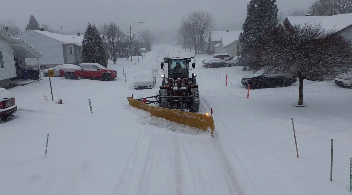 Une déneigeuse dégage une rue résidentielle enneigée bordée de maisons des deux côtés.