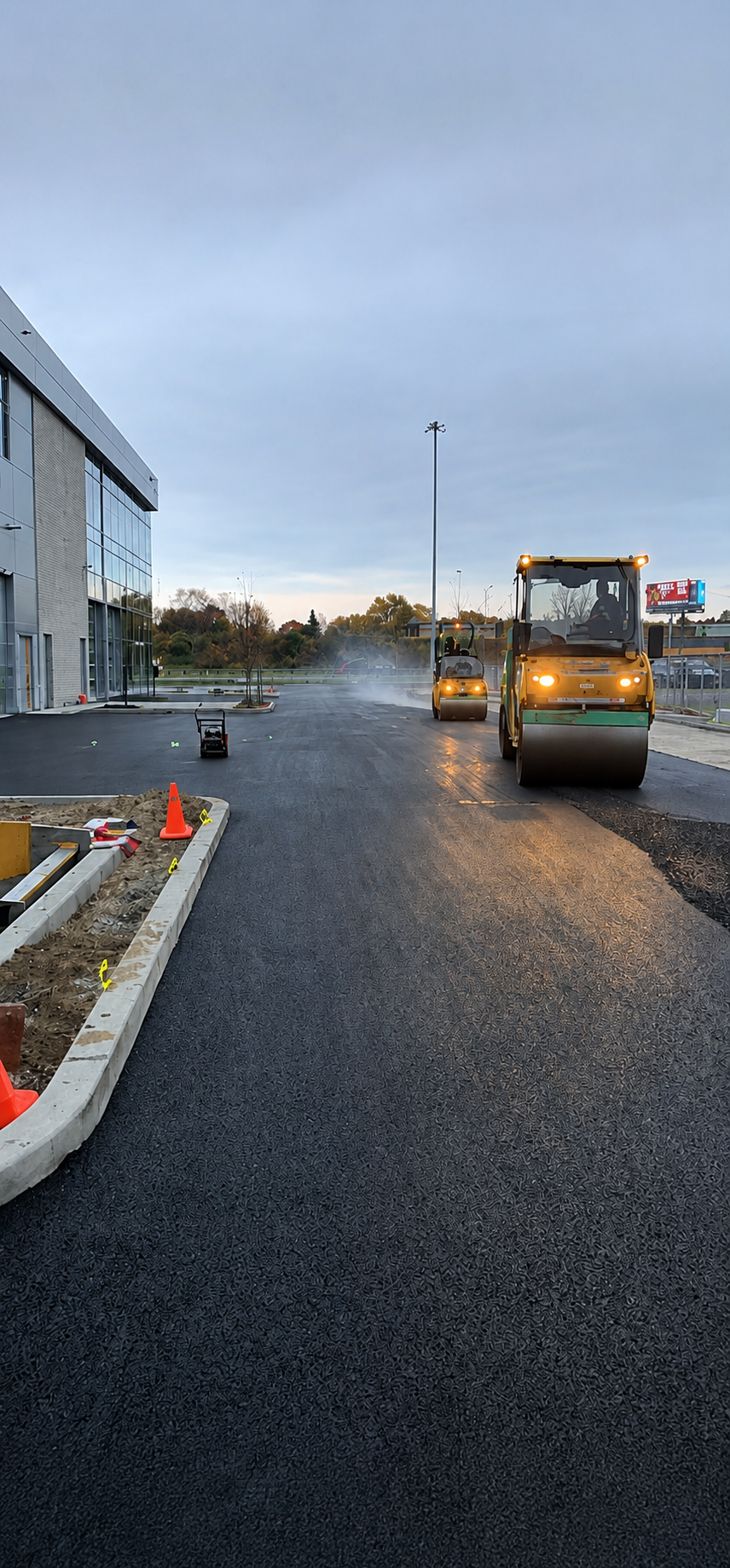 Véhicule de chantier sur chaussée mouillée à côté d'un bâtiment au crépuscule, avec des cônes de signalisation et un ciel nuageux.