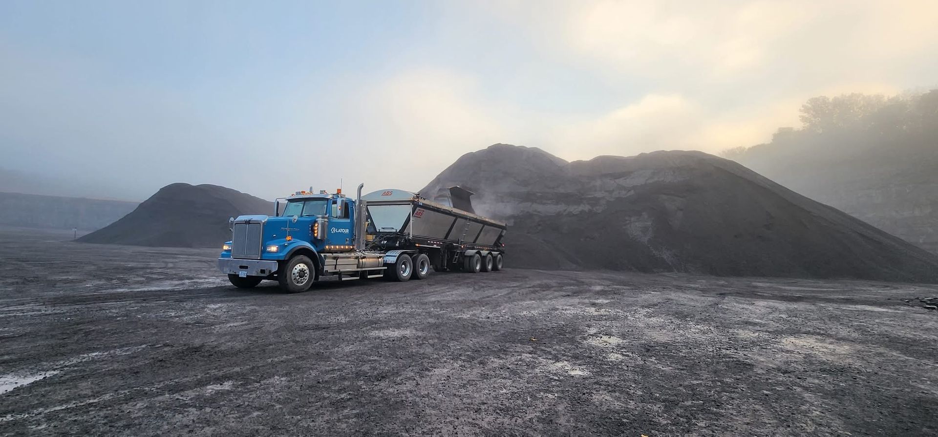 Un camion-benne bleu transporte du gravier noir sur un site minier, avec des tas de roches sous un ciel nuageux.