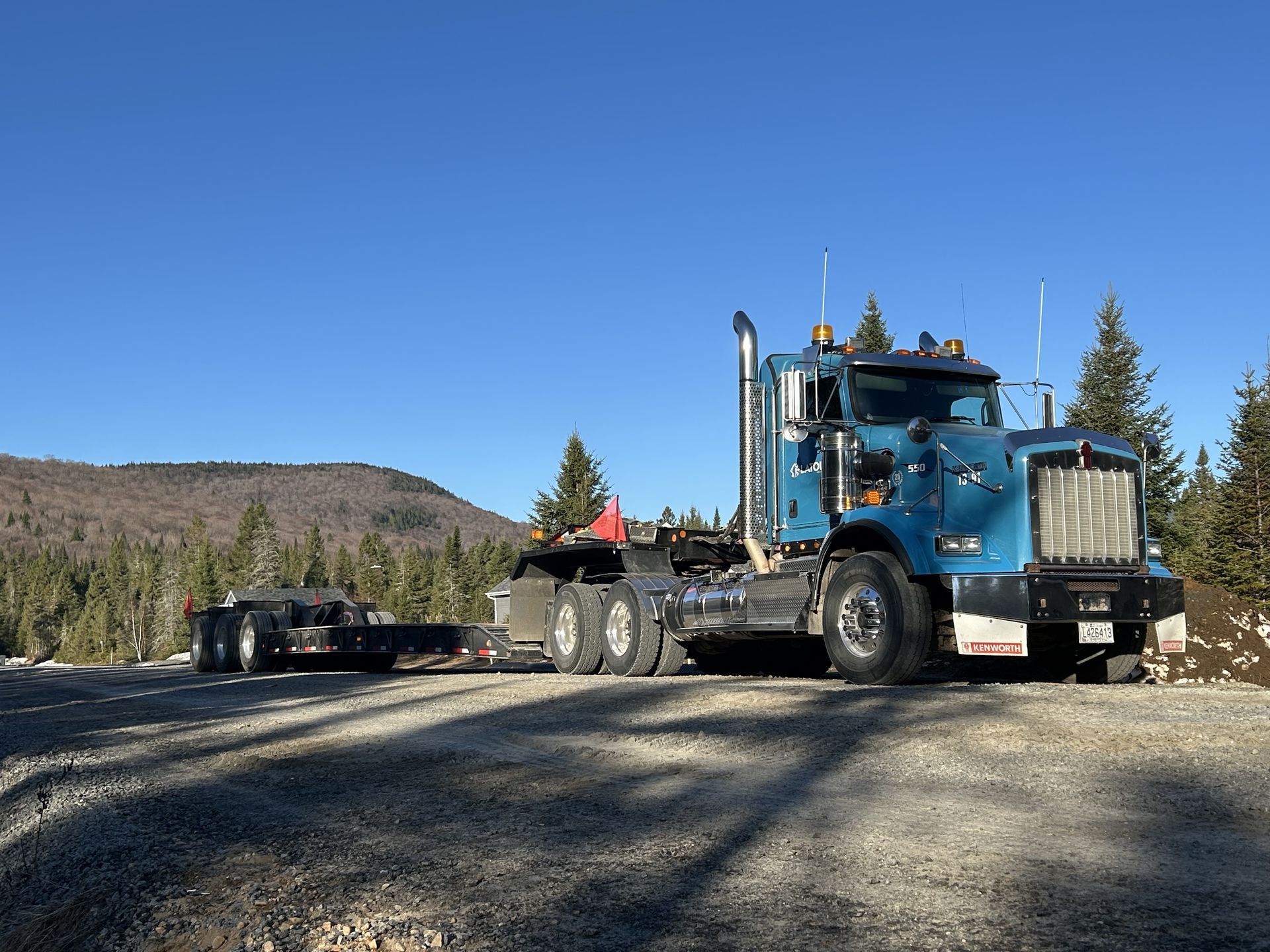 Un semi-remorque bleu sur un chemin de terre, tractant une remorque dans une zone montagneuse boisée, sous un ciel dégagé.
