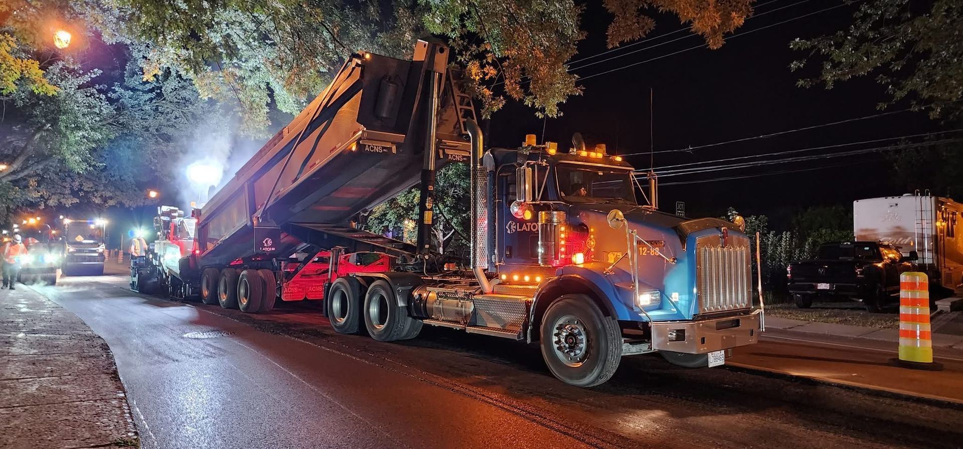 Un gros camion avec une remorque allongée circule de nuit sur une rue mouillée de la ville, les phares se reflétant sur la chaussée.