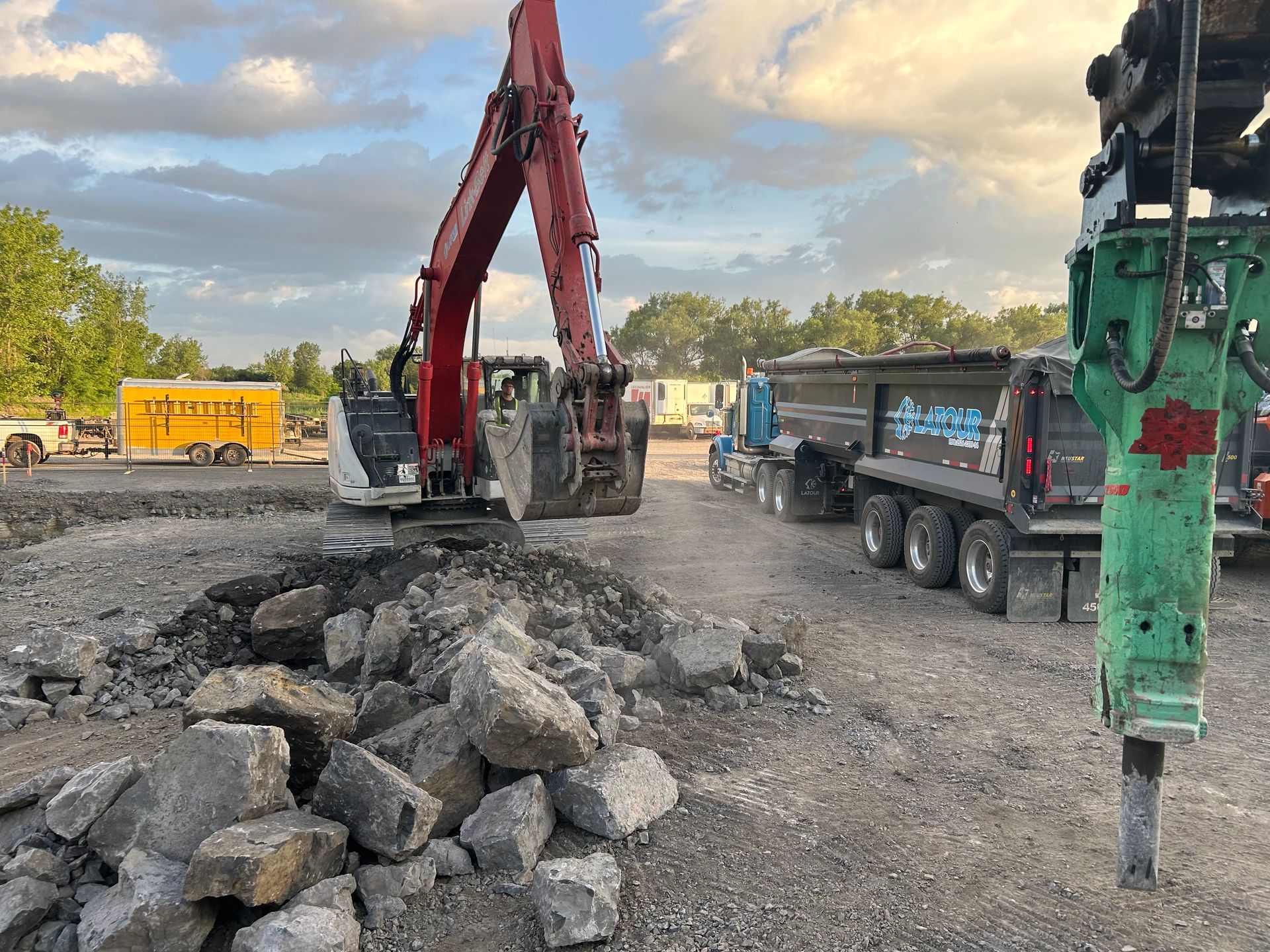 Une excavatrice et des camions-bennes sur un chantier routier, à côté d'un tas de gravats de béton.