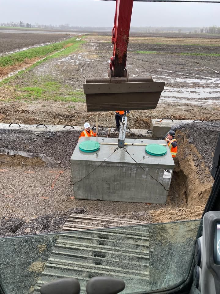 Godet d'une excavatrice déposant un boîtier électrique en béton à côté d'une tranchée boueuse en bord de route, dans un champ rural.