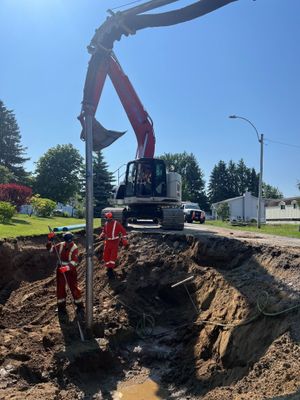 Une équipe de techniciens utilise une grue pour soulever une excavatrice au-dessus d'une grande tranchée en bord de route, sous un ciel bleu.